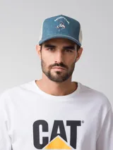 Gorro tipo trucker de color azul denim en la parte frontal y visera, con malla trasera blanca. Presenta un estampado frontal con la inscripción "Caterpillar" y la silueta de un vaquero montando un caballo. Incluye cierre trasero ajustable.