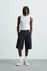 Bermuda de denim rígido color azul oscuro (sin prelavar) con corte amplio (baggy fit). Presenta pliegues frontales, trabillas ajustables en la cintura, bolsillos delanteros y bolsillos traseros de parche, todo resaltado con pespuntes a contraste.