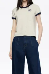 Remera corta de corte ajustado, color beige claro con finas rayas horizontales oscuras. Posee cuello redondo y ribetes en contraste de color azul marino en el cuello y los bordes de las mangas cortas. Lleva un pequeño bordado en forma de corazón con texto en el pecho.