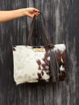 Cartera tipo tote de cuero vacuno con pelo, con estampado de manchas marrones y blancas. Cuenta con doble asa marrón, cierre metálico frontal y un aplique de flecos colgantes de cuero.