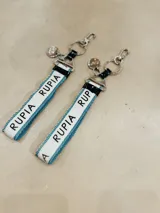 Llavero de cinta tejida con diseño bicolor en blanco y azul, con inscripción de la marca repetida. Incluye herrajes metálicos plateados con mosquetón y argolla para llaves.