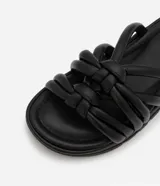 Sandalia plana de color negro con diseño de tiras tubulares acolchadas tipo puffer entrelazadas con nudos. Presenta puntera redonda, plantilla anatómica y suela de goma al tono.