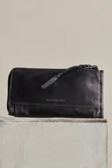 Billetera de cuero color negro con acabado gastado, diseño alargado, cierre perimetral de cremallera y tirador de cuero trenzado. Cuenta con bolsillo exterior con cierre y marca grabada en el frente.