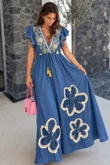 Vestido largo de denim azul con escote en V, mangas cortas con volados y cordón ajustable con borlas en la cintura. Presenta bordados detallados en el pecho y apliques grandes de flores con flecos en la parte inferior de la falda.