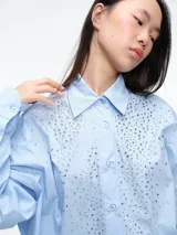 Camisa de corte oversize color celeste, con cuello clásico, cierre frontal de botones y detalle de apliques brillantes en la parte delantera.
