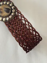 Cinto ancho de cuero trenzado en color marrón, con hebilla circular decorativa que presenta un centro de piedra natural y borde con apliques metálicos esféricos.
