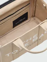 Cartera tipo tote bag de Marc Jacobs, confeccionada en lona color beige con el logo estampado en blanco al frente. Cuenta con doble asa superior, correa de hombro ajustable y extraíble, y cierre superior con cremallera.