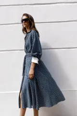 Vestido midi azul marino con rayas blancas, cuello solapa, escote en V y cierre frontal con botones. Cuenta con mangas abullonadas por debajo del codo con puños anchos a contraste, cinturón ajustable del mismo tejido con hebilla metálica y falda evasé.