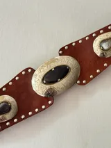 Cinto tipo faja de cuero color chocolate, con apliques metálicos plateados grabados y piedras incrustadas. Presenta detalles de tachas pequeñas en todo el borde y un diseño central ovalado.