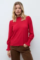 Blusa de color rojo con cuello alto y detalle de volados, mangas largas con puños elásticos en punto smock y textura sutil en la tela.