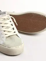 Zapatillas Golden Goose modelo Hi Star, de cuero blanco con estrella dorada con cristales Swarovski, puntera de gamuza gris y refuerzo trasero de cuero negro brillante. La suela es beige con texto manuscrito.