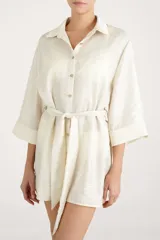 Camisa larga fluida de color crema con cierre frontal de botones, mangas tres cuartos anchas y cinturón ajustable para anudar en la cintura.
