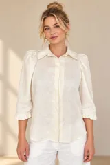 Camisa de lino color blanca, con cuello clásico, mangas tres cuartos abullonadas en los hombros y terminación con volados en los puños, cierre frontal con botones ocultos.