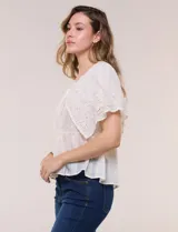 Blusa de estilo romántico color beige confeccionada en algodón, con escote amplio, importante volante capa en broderie calado, cartera frontal con botones tipo perla, bordados verticales calados, cintura marcada con bajo peplum y ruedo recto festonado.