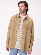 Camisa de gabardina color beige, de corte holgado, con cuello clásico, cierre frontal con botones y dos bolsillos frontales, uno de ellos con parche de la marca.