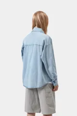 Camisa de denim azul de corte holgado, con cuello clásico, cierre frontal de botones metálicos y bolsillo parche en el pecho con etiqueta cuadrada de la marca.