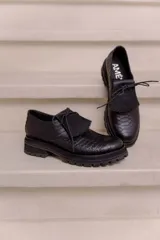 Mocasín de cuero vacuno negro con textura tipo reptil, suela track de goma, cordones finos y solapa con flecos sobre el empeine.