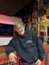 Hoodie negro con capucha y estampa gráfica blanca en la espalda con diseño tipográfico cursivo. Presenta puños y ruedo acanalados.