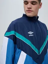 Campera deportiva Umbro modelo Beat, confeccionada en nylon ligero con diseño de bloques de color en blanco, azul marino y verde azulado. Presenta cierre frontal, cuello alto y elásticos en puños y cintura.