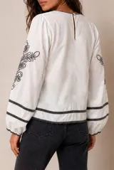 Blusa blanca de manga larga con detalles de bordados florales en tono grisáceo y bandas decorativas en el frente y mangas. Cuenta con cuello redondo y puños elásticos.