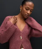 Cardigan de tricot en color borravino, con textura acanalada, escote en V profundo y cierre frontal con un único botón metalizado. Presenta mangas largas y un diseño alargado con amarre ajustable en la espalda.
