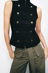 Chaleco negro de cuello subido y manga sisa, con cierre frontal oculto y detalle de botones decorativos metálicos en estilo militar.