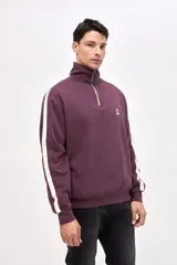 Buzo de felpa con friza, color bordó, con cuello alto y medio cierre metálico frontal. Presenta bandas laterales blancas en las mangas, bolsillos laterales y bordado de la marca en el pecho y la manga.