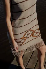 Vestido corto tejido en hilado de algodón color beige con rayas horizontales marrones, cuello de cuero marrón y terminación con flecos. Presenta un diseño geométrico en la parte inferior.