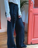 Pantalón de punto color beige, de corte recto y holgado, con cintura elástica y cordón ajustable. Presenta detalles de costuras con flecos verticales a lo largo de las piernas.