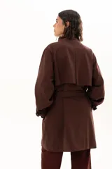 Trench color chocolate con cuello alto, cierre frontal con botones y cinturón ajustable en la cintura. Presenta mangas largas con puños ajustables mediante tiras y hebillas, y bolsillos frontales.