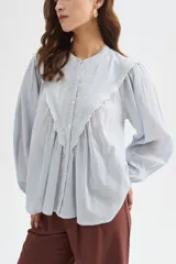Blusa de algodón color beige, con cuello redondo y cierre frontal con botones. Presenta delicados bordados decorativos en tono tostado sobre el pecho y los hombros, mangas largas abullonadas con puños ajustados y diseño holgado.