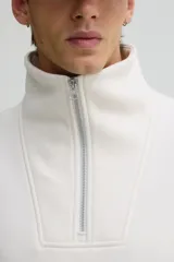 Buzo de tejido pesado color crema, con interior cepillado suave. Presenta cuello elevado, medio cierre metálico y hombros caídos. Los puños y el bajo son de punto acanalado.