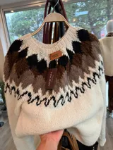 Sweater de punto tejido a mano, con diseño de grecas nórdicas en tonos blanco, marrón y negro en la parte superior. Presenta cuello redondo y mangas largas, con una textura suave y abrigada.