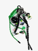 Charm para bolso con diseño lúdico inspirado en los años 90, compuesto por cordones de nylon verde y negro, figuras de personajes, cuentas de acrílico, una cinta métrica pequeña y diversos dijes decorativos.