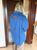 Sobrecamisa de gamuza azul con cierre frontal de botones metálicos dorados, cuello clásico y puños abotonados.