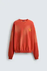 Sudadera de corte relajado, color naranja con efecto tie-dye/deslavado, cuello redondo y manga larga. Presenta un estampado frontal con texto y un trébol de cuatro hojas en amarillo.