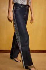 Jeans de tiro bajo y corte recto en denim color azul oscuro. Presenta cinco bolsillos, trabillas para cinturón y cierre frontal con cremallera y botón metálico. Destaca por la aplicación de flecos brillantes en los laterales de ambas piernas.