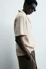 Polo de hombre color crema, de tejido texturado con rayas verticales en relieve, corte relajado, cuello bowling y cierre frontal parcial con botones.