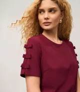 Blusa de manga corta color bordó, con cuello redondo y apliques de lazos decorativos apilados en las mangas.