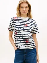 Remera Tommy Hilfiger de corte regular, confeccionada en punto interlock de algodón blanco con rayas horizontales azul marino y diseño floral gráfico superpuesto con detalles en rojo. Presenta cuello redondo y mangas cortas.
