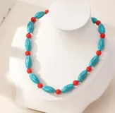 Collar de cuentas asimétrico compuesto por piezas alargadas de imitación piedra turquesa con vetas negras, intercaladas con pequeñas cuentas esféricas de color rojo. Incluye cadena extensora metálica en tono dorado.