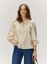 Camisa de algodón color beige con patrón texturizado en relieve, escote en V con cuello mao, mangas largas abullonadas con puños ajustados y detalles de volados en los hombros. Cierre frontal con botones.