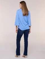 Blusa celeste de tejido texturado con relieve floral, escote redondo con muesca central, mangas tres cuartos estilo campana y ruedo curvo.