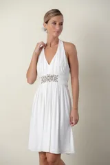 Vestido corto de color blanco con escote halter y espalda descubierta. Presenta un diseño drapeado en el busto y una banda en la cintura decorada con bordados y pedrería. La falda tiene un corte fluido con movimiento.
