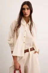 Camisa de algodón color crudo, con calce relajado, cuello clásico, botones delanteros, mangas largas con sutil volumen y ruedo asimétrico.