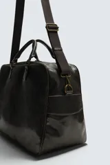 Bolso de viaje de diseño rectangular con acabado craquelado en tono oscuro. Cuenta con doble asa de mano, compartimento principal con cierre de cremallera, bolsillo interior con cierre y correa de hombro ajustable y extraíble.