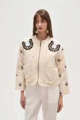Campera estilo bomber color beige claro, con cuello redondo, cierre frontal y detalles de volados en los bordes. Presenta bordados florales en negro y blanco en la zona del pecho y bordados de tréboles negros en las mangas.