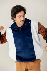 Buzo de textura afelpada con diseño de bloques de color en azul, blanco y marrón. Presenta cuello alto con cierre frontal parcial mediante botones a presión.