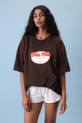 Remera oversize color lila con estampa frontal gráfica en tonos marrón y crema. Presenta cuello redondo, mangas cortas y un corte holgado.