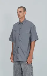 Camisa de manga corta en color gris, confeccionada en popelina de algodón. Presenta un corte oversize, cuello clásico, cierre frontal con botones y dos bolsillos delanteros, uno de ellos con solapa.